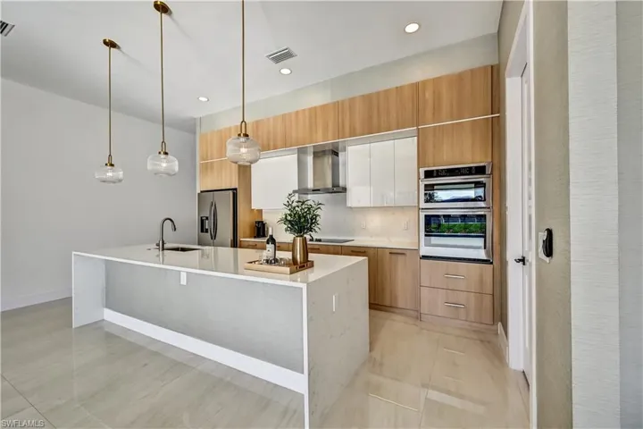 Dual tone kitchen featuring decorative light fixtures, two tone color scheme, stainless steel appliances, a center island with sink, and light stone counters