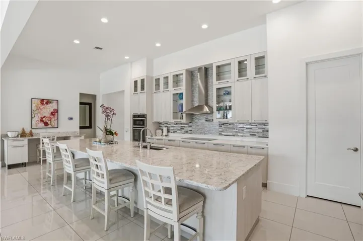 Spacious kitchen featuring a large island with a surface-mounted sink, built-in double ovens, a contemporary range hood, a tiled backsplash, and recessed lighting