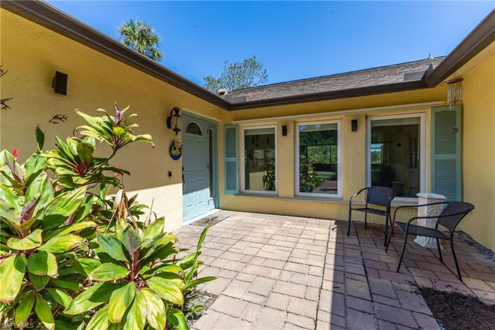 Paver patio entry featuring a light blue door with an arched window, stucco exterior, multiple large windows with light blue shutters, and mature landscaping