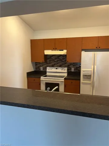 Kitchen with under cabinet range hood, white appliances, a peninsula, dark countertops, and tasteful backsplash