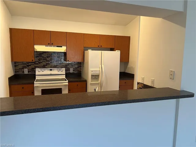 Kitchen featuring under cabinet range hood, white appliances, a peninsula, backsplash, and dark countertops