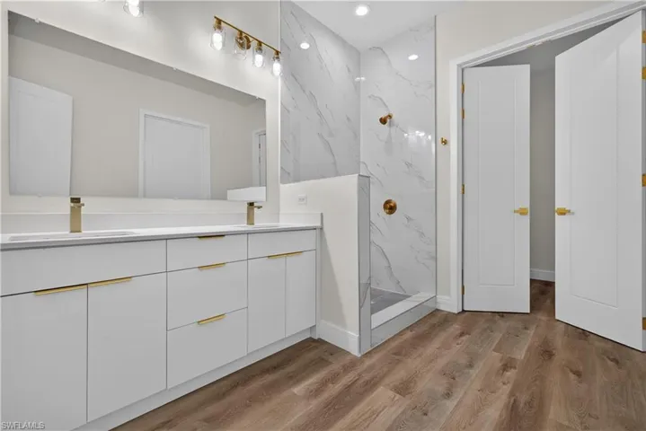 Dual-sink vanity with white cabinetry, brass hardware, wood-finish flooring, walk-in shower with stone-patterned tile surround, and expansive wall-mounted mirror