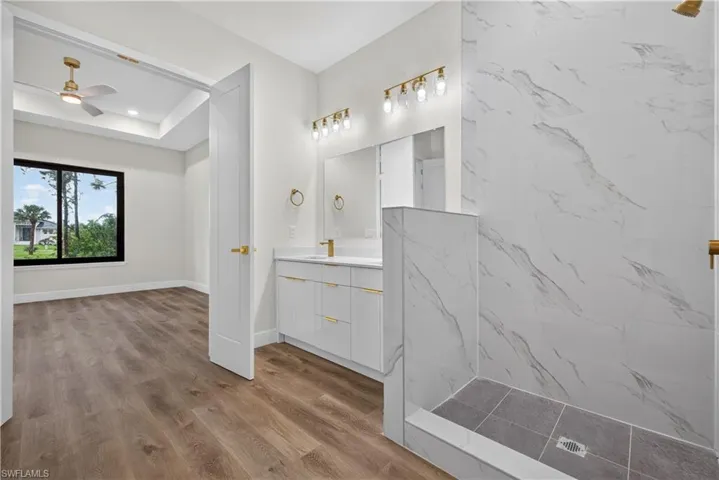 Bathroom featuring a walk-in shower with large-format wall tile, white vanity cabinetry with gold-tone hardware, wood-finish flooring, and multi-bulb vanity lighting