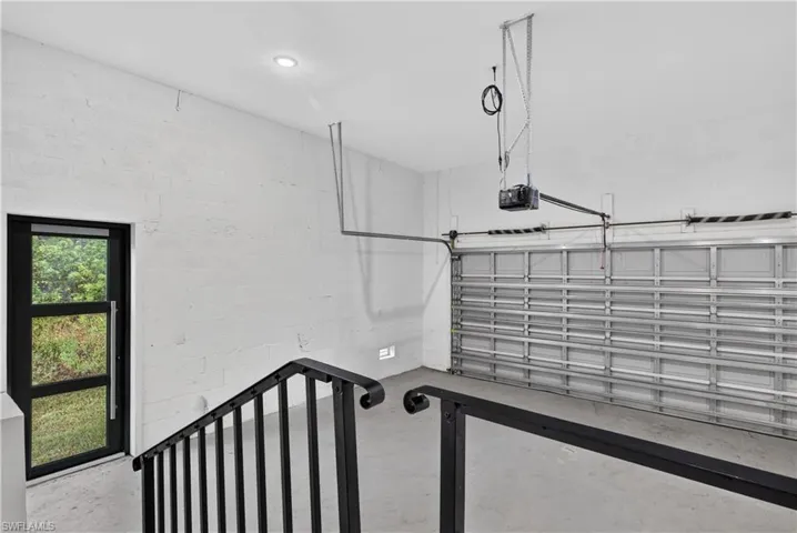 Garage space featuring white painted masonry walls, a sectional metal overhead door, recessed ceiling lighting, a dedicated exterior pedestrian door with glass panels, and a matte black metal stair railing
