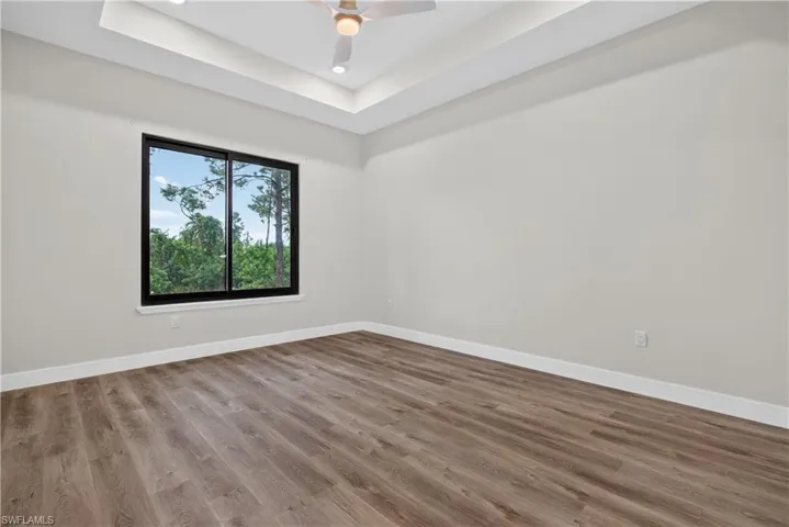 Tray ceiling, wood-finish flooring, ceiling fan, and large window with exterior greenery