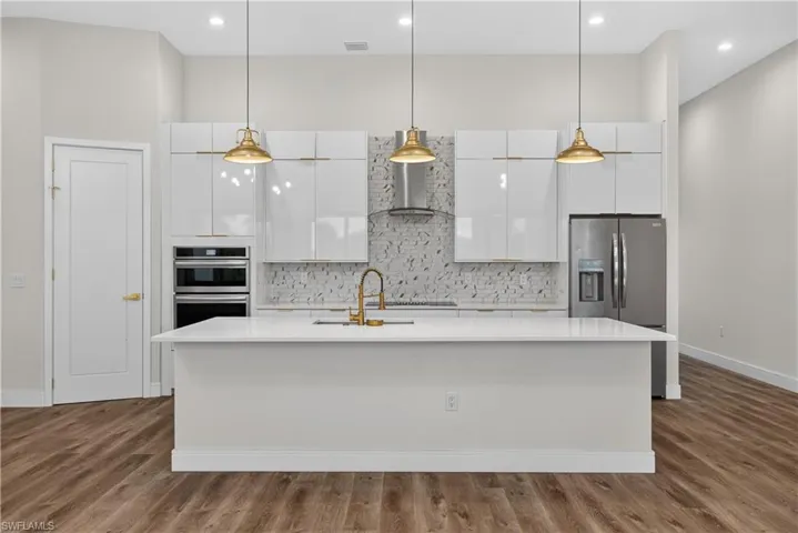 Kitchen featuring white high-gloss cabinetry, large center island with a quartz countertop, stainless steel appliances, patterned tile backsplash, and hanging pendant lighting over wood-finish flooring