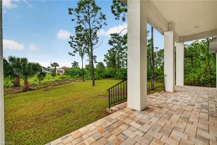 Covered patio featuring paver flooring, structural pillars, and iron handrails, overlooking a grass yard with mature landscaping