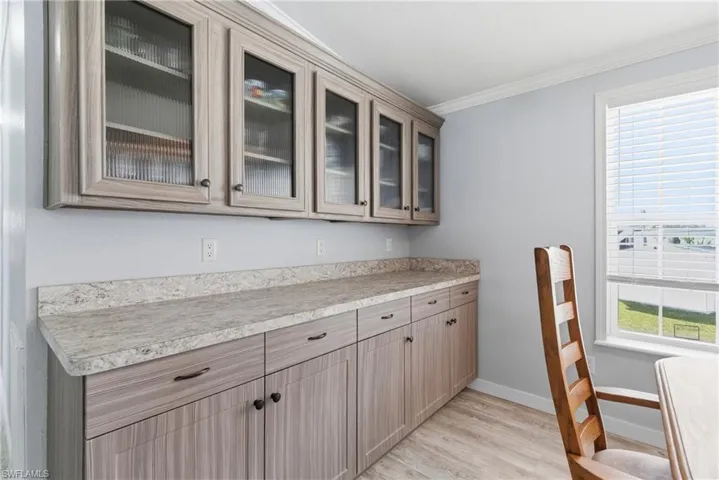 Bar featuring light countertops, glass insert cabinets, ornamental molding, light wood-style flooring, and light brown cabinetry