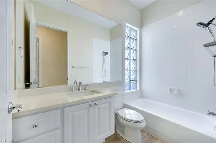 Bathroom featuring a white vanity with a light-colored countertop and integrated sink, a large wall-mounted mirror, a bathtub with a shower fixture, a toilet, and a glass block window
