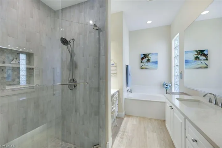Upgraded shower with glass enclosure, light-toned wood-finish flooring, built-in tub, dual vanity with undermount sinks, and a privacy window featuring glass blocks