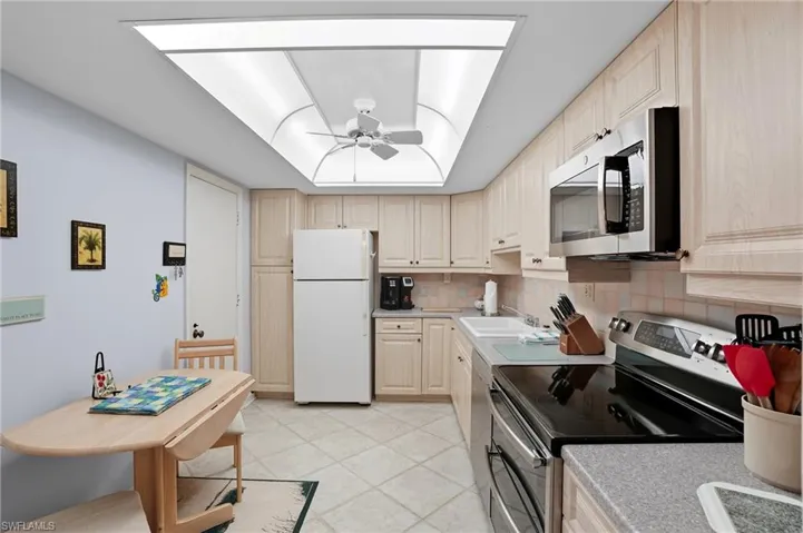 Kitchen featuring light wood-finish cabinetry, neutral tile flooring, and a luminous ceiling panel with a ceiling fan