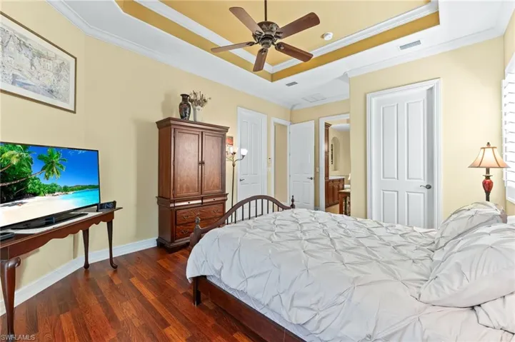 Bedroom with wood flooring, a raised ceiling, ceiling fan, ornamental molding, and connected bathroom