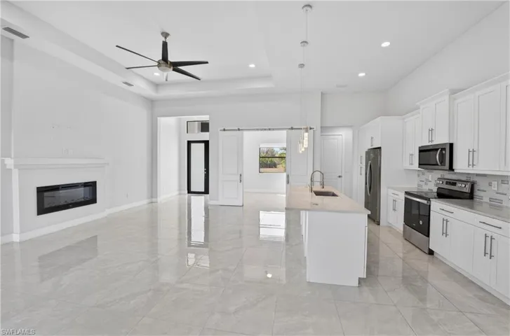 Kitchen with stainless steel appliances, white cabinetry, decorative light fixtures, an island with sink, and a glass covered fireplace
