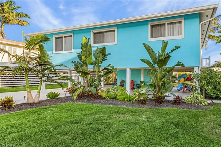 Turquoise-painted elevated coastal residence featuring multiple windows, a well-maintained lawn, lush tropical landscaping with palm trees, and a paved driveway