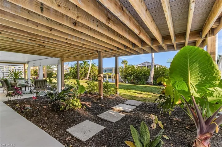 Under-deck patio with concrete flooring, accented by a landscaped garden bed with stepping stones, mature tropical foliage, and an expansive lawn