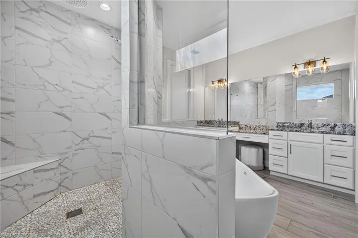Spacious bathroom featuring a freestanding tub, dual vanities with stone countertops, a large walk-in shower with a built-in bench, and wood-finish flooring