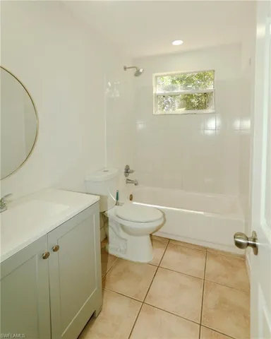 Full bathroom featuring a white tub and shower combination with subway tile surround, a window providing natural illumination, a vanity with a white countertop and gray cabinetry, a circular mirror, and neutral-toned floor tiling