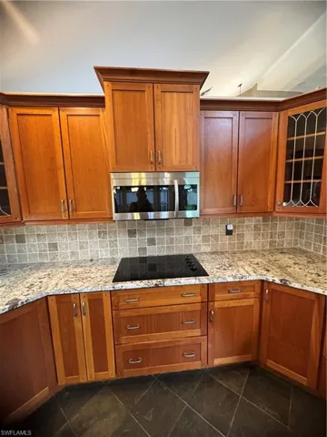 Kitchen featuring decorative backsplash, stainless steel microwave, cooktop, glass insert cabinets, and light stone counters