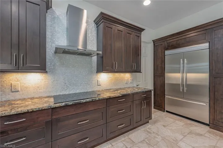 Kitchen with dark brown cabinets, stainless steel built in refrigerator, wall chimney range hood, dark stone counters, and tasteful backsplash