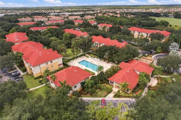 Community featuring multiple buildings with red tile roofs, a central swimming pool with lounge chairs, mature trees, and paved parking areas