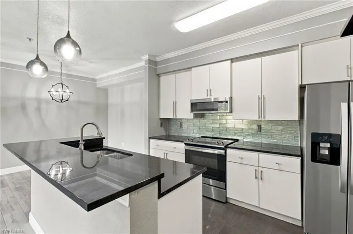 Kitchen featuring an island with an undermount sink, stainless steel appliances, white cabinetry, dark countertops, and a tiled backsplash