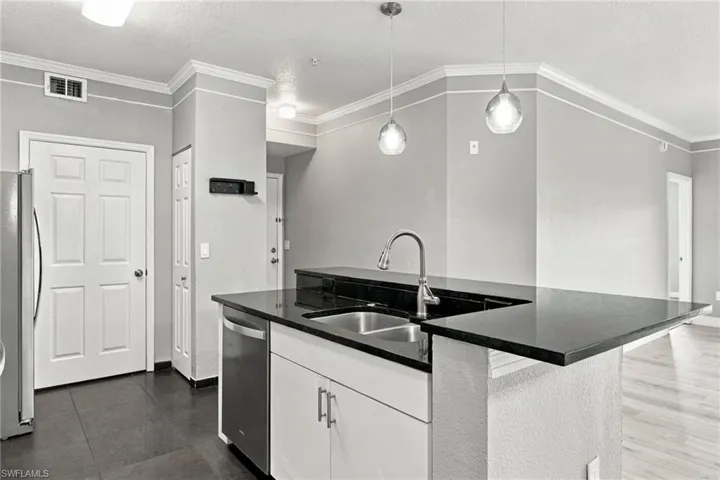 Kitchen island with black countertop and integrated stainless steel sink, stainless steel dishwasher, white cabinetry with silver hardware, and pendant lighting