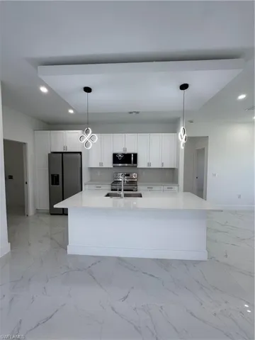 Kitchen with white cabinetry, stainless steel appliances, light stone countertops, a center island with sink, and decorative light fixtures