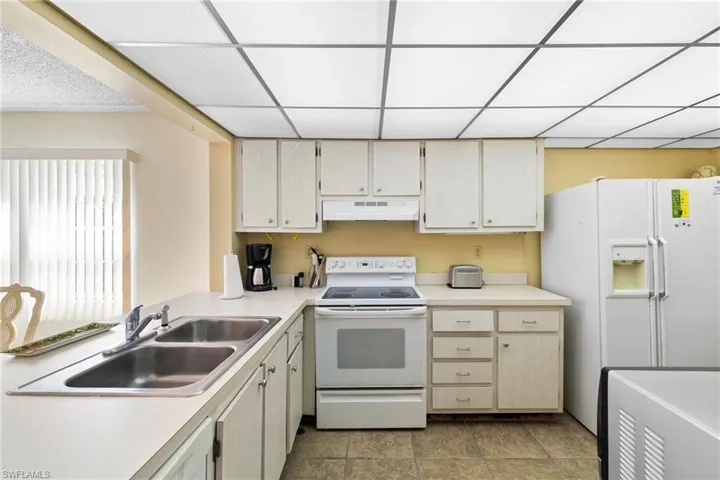 Kitchen with white appliances, light countertops, under cabinet range hood, a drop ceiling, and light tile patterned floors