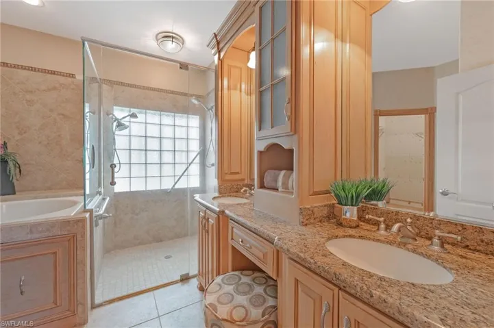 Bathroom featuring a dual vanity with granite countertops and undermount sinks, a built-in makeup area, extensive wood cabinetry, a glass-enclosed shower with multiple showerheads, and a built-in bathtub with tiled surround