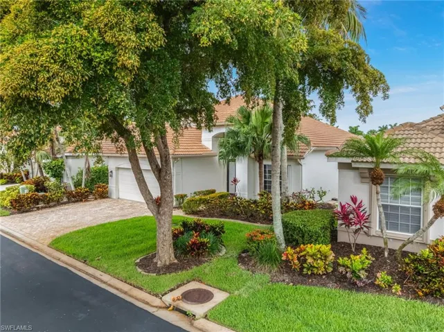 Tile roof residence with a paver driveway, an integrated garage, and a lush front yard featuring mature trees, shrubs, and colorful plantings