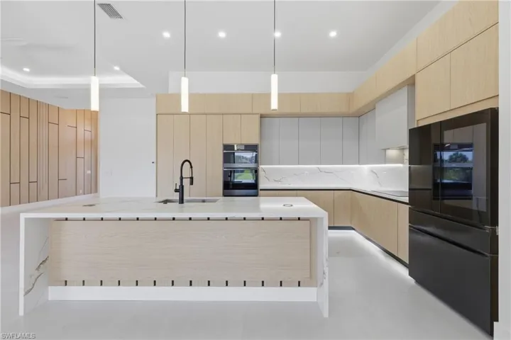 Dual tone kitchen featuring a kitchen island with sink, black appliances, hanging light fixtures, light stone countertops, and modern cabinets