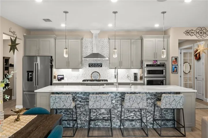 Kitchen featuring stainless steel appliances, wall chimney range hood, a center island with sink, tasteful backsplash, and light wood-look tile flooring