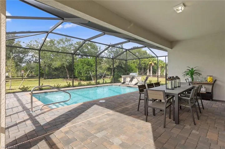 Screened-in outdoor living space featuring a rectangular pool with handrail, paver decking, an outdoor kitchen with a built-in grill, and a covered patio area with an overhead light fixture