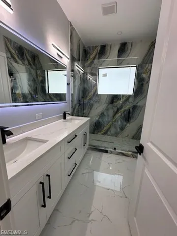 Bathroom featuring double vanity, a marble finish shower, and light marble finish flooring