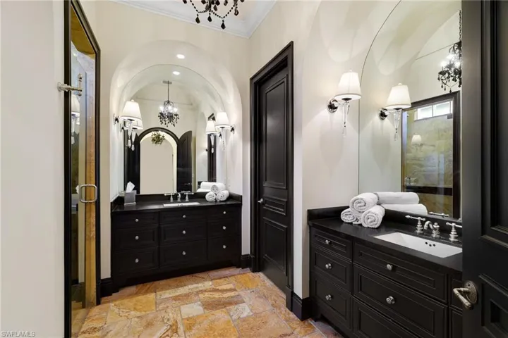 Bathroom with a chandelier, a shower stall, two vanities, stone tile floors, and crown molding