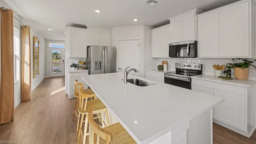 Modern kitchen featuring a large central island with an undermount sink, white cabinetry, stainless steel appliances, white subway tile backsplash, and wood-finish flooring