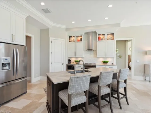 Kitchen with wall chimney range hood, decorative backsplash, light stone countertops, stainless steel fridge, and a kitchen island with sink