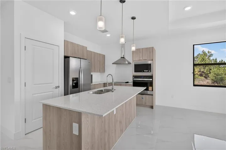 Modern kitchen featuring a large island with a light-toned countertop, stainless steel appliances, wood-finish cabinetry, and contemporary pendant lighting