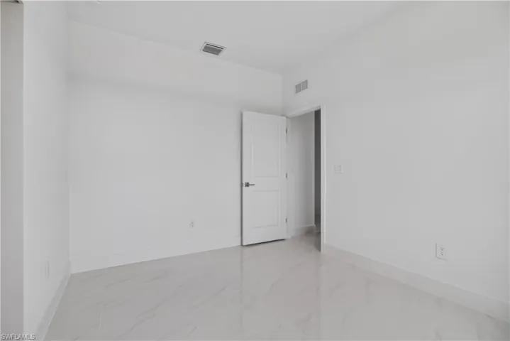 Spacious room featuring light-toned tile flooring, clean white walls, a single paneled door, and a ceiling-mounted ventilation grate