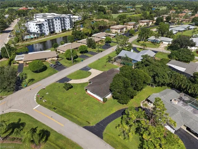 Aerial perspective featuring a single-story residence with a shingled roof, corner lot placement, curved asphalt driveway, and mature landscaping