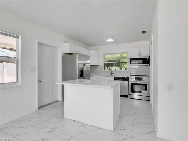 Modern kitchen featuring white shaker cabinetry, stainless steel appliances, a center island with a stone countertop, white tiled flooring, and a large window providing ambient light