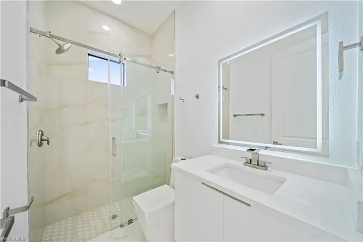 Modern bathroom featuring a single vanity with a white countertop and integrated sink, a frameless backlit mirror, and a glass-enclosed shower
