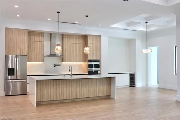 Kitchen featuring light brown cabinetry, stainless steel appliances, modern cabinets, wall chimney range hood, and hanging light fixtures