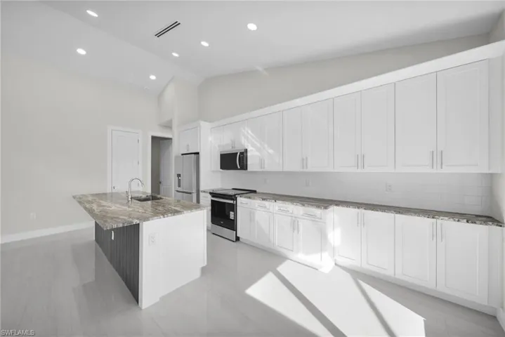 Kitchen featuring an island with sink, stainless steel appliances, decorative backsplash, white cabinetry, and light stone counters
