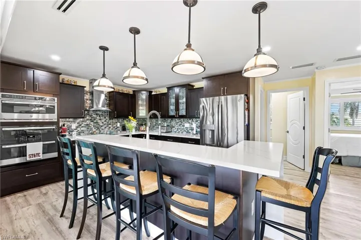 L-shaped kitchen bar island with waterfall quartz counters, glass insert cabinets, dark brown cabinetry, appliances with stainless steel finishes, and recessed lighting