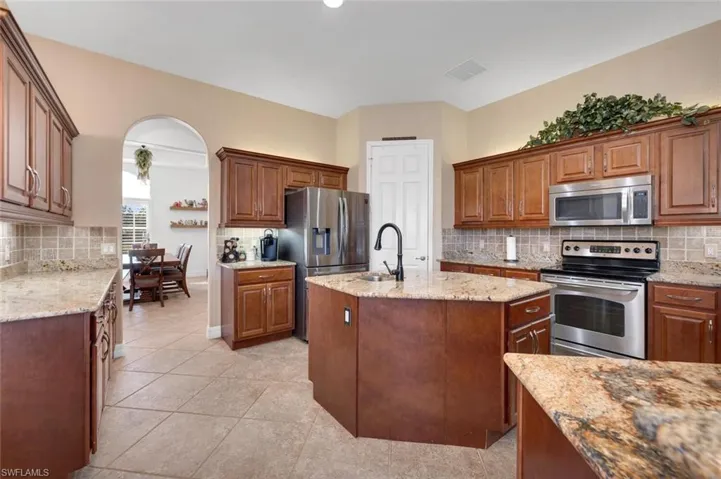 Kitchen with backsplash, light stone counters, stainless steel appliances, a kitchen island with sink, and light tile patterned floors