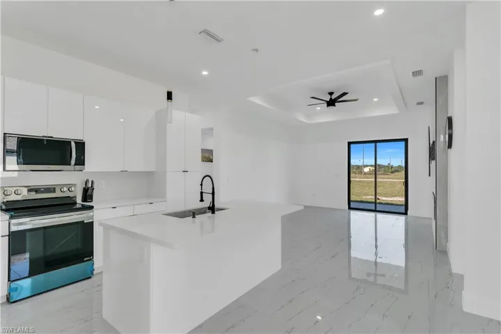 Kitchen with stainless steel appliances, a raised ceiling, an island with sink, white cabinetry, and light marble finish floors