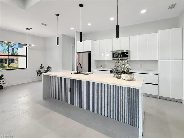 Contemporary kitchen featuring a large island with fluted paneling and light-toned countertop, white high-gloss cabinetry, stainless steel appliances, and sleek pendant lighting