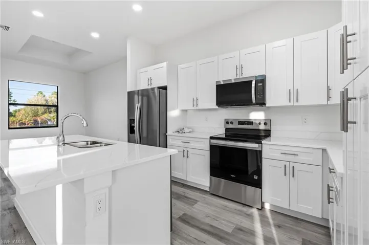 Kitchen featuring stainless steel appliances, a center island with sink, light stone countertops, white cabinets, and light wood-type flooring