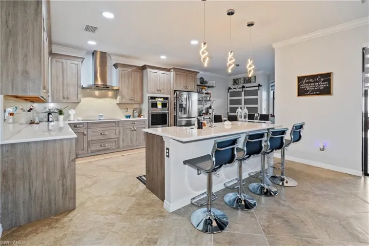 Kitchen with light stone countertops, a kitchen bar, crown molding, wall chimney range hood, and hanging light fixtures